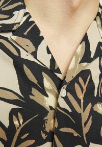 Chemise avec col arborant un motif botanique noir et beige, ornée de boutons crème et d'une texture de tissu lisse. Motifs de feuilles remarquables tout au long.