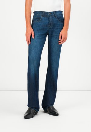 Person wearing dark blue straight-leg jeans and black pointed-toe shoes, standing against a plain white background.