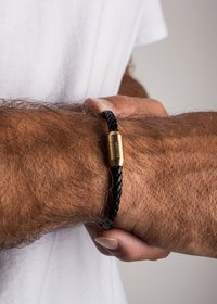 Black braided leather bracelet with a gold-tone cylindrical clasp. Textured surface and secure fit on a wrist. Simple and functional design.