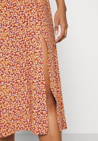 Jupe midi fleurie orange avec une fente sur le côté. Présente un motif de fleurs blanches et violettes sur un tissu texturé.