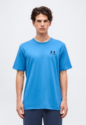 Fiatal férfi kék Under Armour pólóban, egyszerű világos háttér előtt áll, semleges arckifejezéssel előre néz.