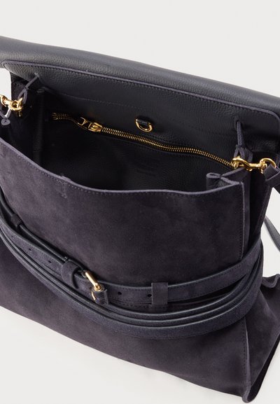Sac bandoulière en daim gris foncé avec rabat avant, sangle réglable et poche zippée dorée à l’intérieur du compartiment principal.