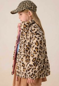 Jonge meid in een fleece jasje met luipaardprint en een bijpassende luipaardprint pet, staand in profiel tegen een effen achtergrond.