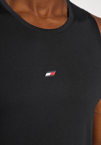 Gros plan d'une personne portant un haut noir sans manches avec un petit logo comportant des rectangles blancs, rouges et bleu marine sur la poitrine.