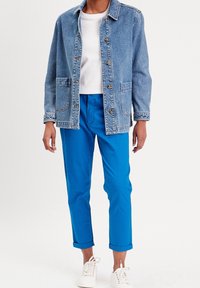 Jeansjacke in Hellblau mit zwei Fronttaschen und goldenen Details, kombiniert mit leuchtend blauen, hochgekrempelten Hosen und weißen Sneakers.