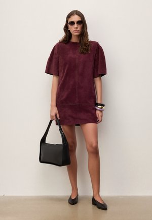 Sieviete, kas valkā īsām piedurknēm apsarkanu kleitu, melnas saulesbrilles, melnus zempapēžu apavus, nes melnu rokassomu un valkā aproces.