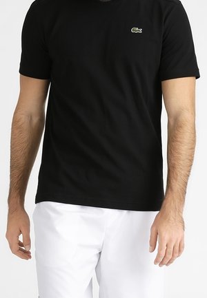 Man draagt een effen zwart T-shirt met korte mouwen met een klein groen krokodillenlogo op de borst en witte broek.