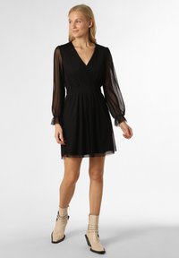 Schwarzes Kleid mit Wickeldesign, durchsichtigen langen Ärmeln und tailliertem Schnitt. Getragen mit beigen Knöchelstiefeln mit einem einzigartigen Riemendetail.