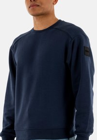 Felpa blu navy realizzata in tessuto morbido, con scollo a girocollo e maniche raglan. Include un'etichetta con logo nero sulla spalla sinistra.