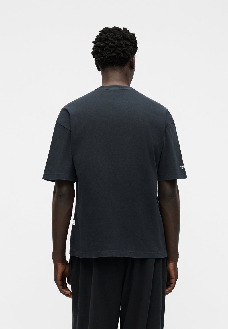 Mand med kort krøllet hår iført en mørk oversized T-shirt og løse bukser, set bagfra mod en ensfarvet lys baggrund.