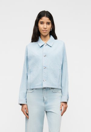 Femme aux longs cheveux bruns portant une veste en jean bleu clair boutonnée et un jean taille haute assorti, debout devant un fond uni.