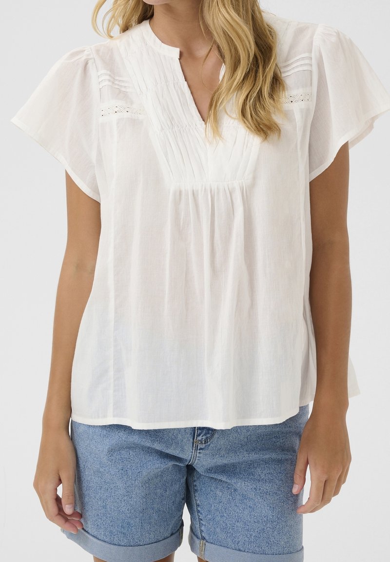 Blouse blanche à manches courtes avec un design plissé et des détails en dentelle, dotée d'un col mandarin et d'une texture légère. Portée avec un short en jean.