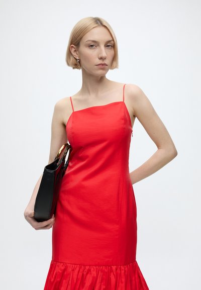 Femme aux cheveux blonds courts portant une robe rouge sans manches, tenant un sac à main noir avec des poignées dorées sur un fond uni.