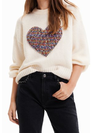 Femme portant un pull en maille crème avec un grand cœur tricoté multicolore sur la poitrine, associé à un jean noir taille haute.