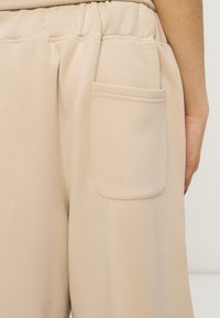 Μπεζ ελαστικό παντελόνι με λάστιχο στη μέση και πίσω τσέπη στη δεξιά πλευρά, σε κοντινή λήψη από τη μέση μέχρι το μέσο του μηρού.