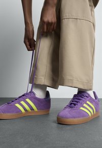 Személy bézs nadrágot és fehér zoknit viselve lilás Adidas sportcipőt köt meg, amelyen neon sárga csíkok és gumi talpak találhatók, mindez egy szürke padlón.
