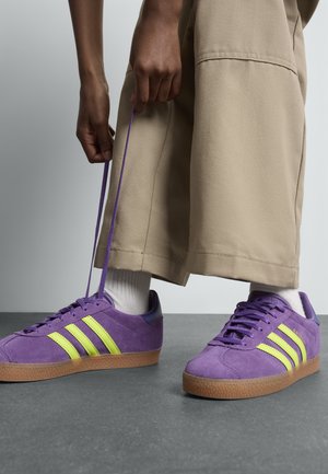 En person som bär beige byxor och vita strumpor knyter lila Adidas-sneakers med neongula ränder och gummisulor på ett grått golv.