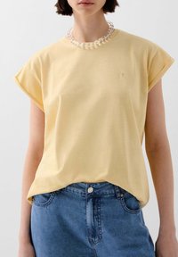 Person, der ein blassgelbes T-Shirt mit hochgerollten Ärmeln, blaue Jeans und eine klare, runde Perlenkette trägt, bis zur Mitte des Gesichts zugeschnitten.