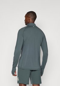 Μακρυμάνικη αθλητική μπλούζα σε ήσυχο πράσινο ύφασμα, με ψηλό γιακά και οπές για τους αντίχειρες, σε συνδυασμό με αντίστοιχες βερμούδες. Απλός σχεδιασμός, χωρίς σχέδια.