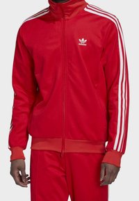 Red athletic jacket made of smooth fabric, featuring a high collar, full zipper, and white vertical stripes on the sleeves. adidas logo on chest.
