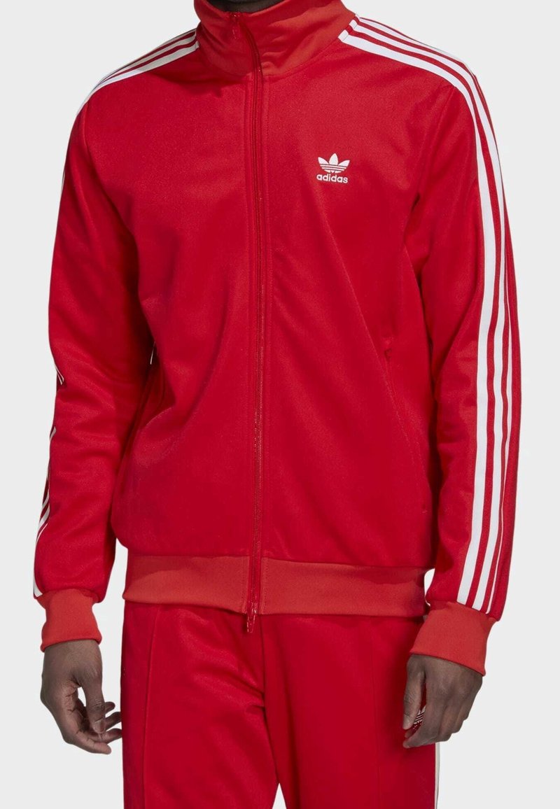 Red athletic jacket made of smooth fabric, featuring a high collar, full zipper, and white vertical stripes on the sleeves. adidas logo on chest.