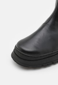 Čierna kožená členková čižma s hladkým povrchom, okrúhlou špičkou, gumovou podrážkou a textúrovaným detailom pozdĺž spodného okraja.