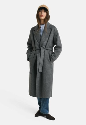 Grijze wollen blend lange jas met een ceintuur in de taille, reverskraag en zijzakken, gecombineerd met donker jeans en schoenen. Een neutrale pet maakt de look compleet.