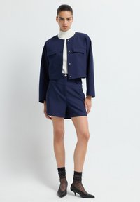 Vrouw staat in een marineblauwe cropped jas en bijpassende hoge shorts, witte turtleneck, doorschijnende zwarte sokken en zwarte kitten heels.