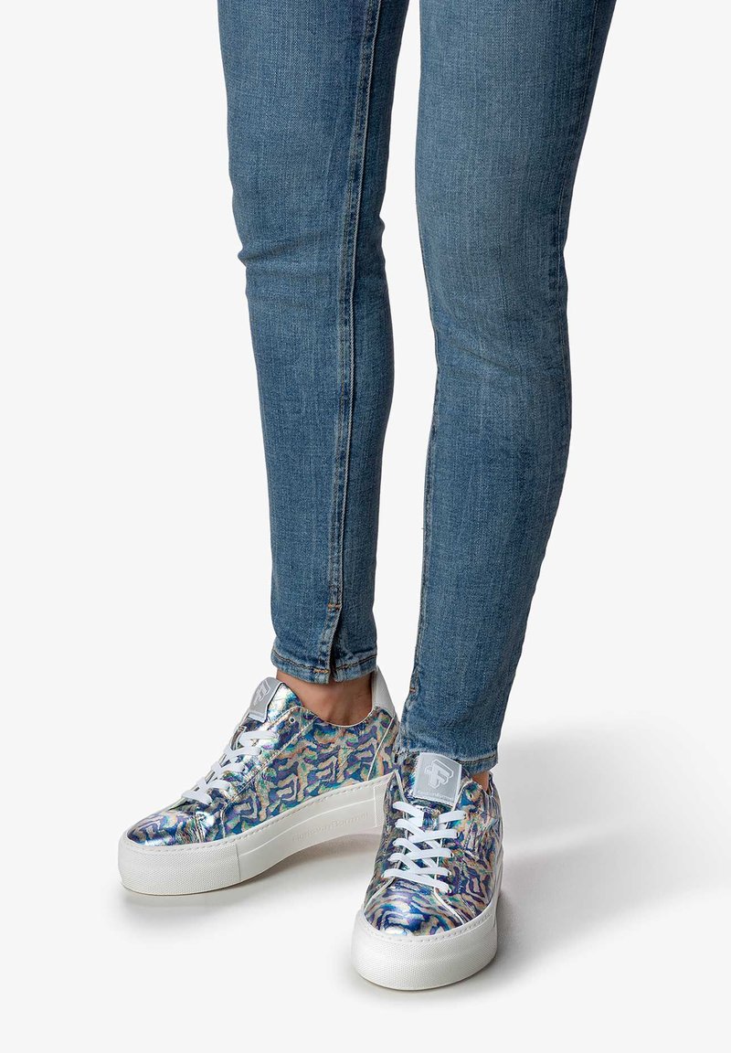 Bunt gemusterte Sneakers mit einem glänzenden Obermaterial, weißen Schnürsenkeln und einer dicken weißen Sohle, kombiniert mit engen blauen Jeans.