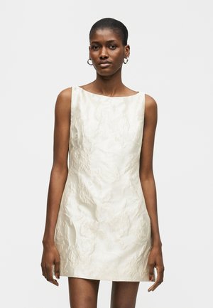 Γυναίκα με μίνι φόρεμα χωρίς μανίκια, ελεφαντόδοντο, με υφή και λαιμόκοψη καραβόπανο, στέκεται μπροστά σε απλό ανοιχτό γκρι φόντο.