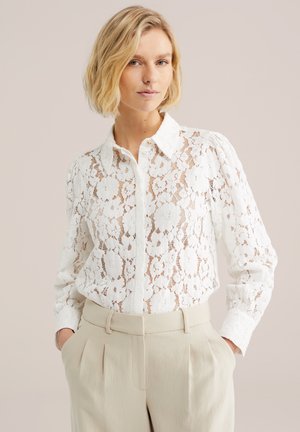 Femeie blondă purtând o bluză albă cu dantelă florală și mâneci lungi, introdusă în pantaloni bej plisati, cu talie înaltă, stând cu mâinile în buzunare.