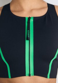 Čierny športový crop top s predným zipsom, ktorý má jasne zelené akcenty pozdĺž švov a logovú nášivku blízko zipsu.