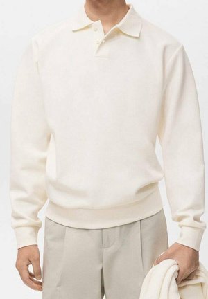 Mann trägt ein cremefarbenes, langärmeliges Poloshirt und beige Hosen, hält einen zusammengefalteten cremefarbenen Pullover in der rechten Hand.