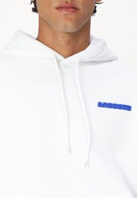 Felpa bianca con cappuccio a coulisse, dotata di una patch del logo blu sul lato sinistro del petto. Il tessuto sembra morbido e presenta un design minimalista.