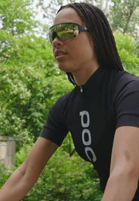 Jersey de ciclismo negro con el logo "POC", gafas de sol reflectantes, cabello trenzado, en un entorno al aire libre con follaje verde de fondo.