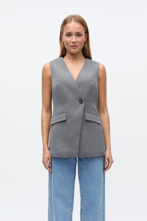 Blazer sans manches gris avec un col en V, fermeture à un bouton et deux poches avant. Porté avec un jean bleu clair à jambes larges.