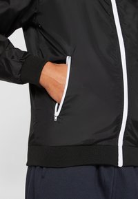 Zwarte jas met een gladde textuur, voorzien van witte ritsdetails en elastische manchetten. Inclusief zijzakken met ritssluitingen.