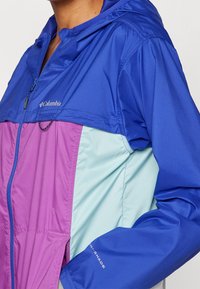 Colorful jacket featuring a blue hood and upper body, purple midsection, and teal lower section. Made from lightweight, breathable fabric.