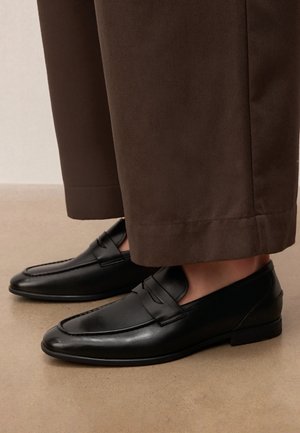 Czarne skórzane mokasyny noszone z brązowymi spodniami o szerokich nogawkach, stojące na gładkiej beżowej podłodze.