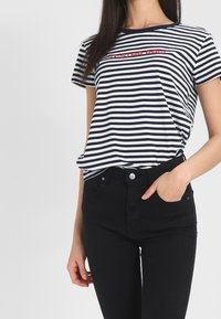 Camiseta de algodón con rayas verticales en navy y blanco, escote redondo y detalle en texto rojo en el pecho. Combinada con jeans negros.