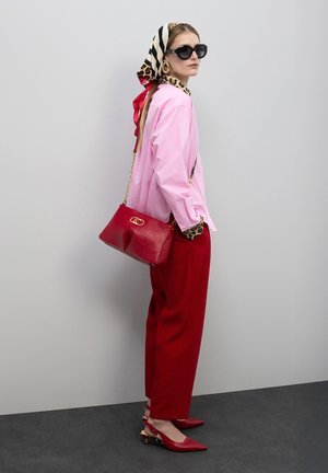 Bolso de cuero rojo con herrajes dorados, usado como bandolera. Modelo con una camisa rosa, pantalones rojos, gafas de sol negras y un pañuelo de cabeza estampado.