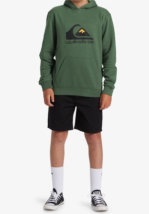 Enfant portant un sweat à capuche vert Quiksilver, un short noir, des chaussettes blanches montantes et des baskets Converse noires, debout devant un fond blanc.