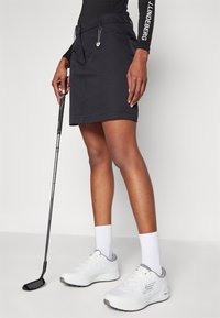 Žena v černé golfové sukni držící putter, obutá v bílých teniskách a ponožkách, stojící před jednoduchým světlým pozadím.