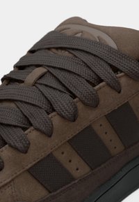 Κοντινό πλάνο καφέ σουέντ αθλητικού παπουτσιού με παχιά σκουρό καφέ κορδόνια και μαύρες υφασμάτινες ρίγες στο πλάι.