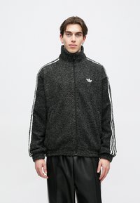 Φλις μπουφάν με φερμουάρ σε σκούρο γκρι χρώμα με ψηλό γιακά, διαθέτει λευκές ρίγες στα πλάγια και κεντημένο λογότυπο στο στήθος, ανθεκτική υφή.