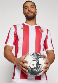 Rød og hvitstripet fotballdrakt med Kappa-logo i gull på skuldrene. Spilleren holder en svart og hvit mønstret fotball.
