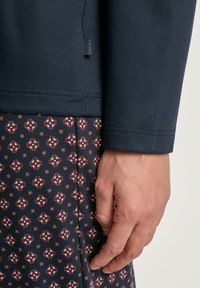 Marineblauwe lange mouwen top met geribbelde manchetten, gecombineerd met een donkere stof met een patroon van diamantvormen in rood en wit. Etiket zichtbaar.