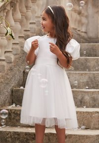 Ung pige i hvid kjole og perlehårbånd står på stentrapper, smiler og er omgivet af svævende sæbebobler.