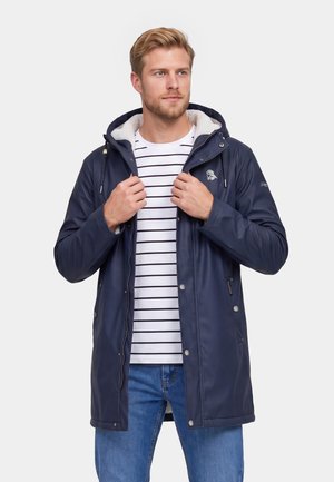 Man draagt een marineblauw capuchonjack over een wit-zwart gestreept shirt, past de rits van het jack aan, staand tegen een effen achtergrond.