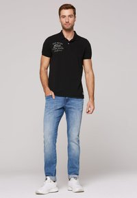 Polo nero a maniche corte con colletto, dotato di tasca frontale e testo stampato. Abbinato a jeans azzurri e sneakers bianche.